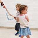 Two young children with curly hair walk side by side; one in a white tank top and denim skirt, the other in a plaid shirt and shorts. Both wear the Child Safety Wristband, featuring an anti-slip Velcro design for 360-degree free rotation, as they stroll on the light-colored floor.