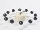 The Clock, background wall, wall clock features a modern European design with a cylindrical white base and twelve metal rods ending in black spheres for the hours. Minimalist black hour and minute hands enhance its striking appearance in any interior space.