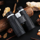 A high resolution binocular telescope with a silver focus knob sits on a wooden log, demonstrating its powerful magnification. Water droplets suggest recent use or rain, while surrounding rocks, pine cones, and dried plants create an ideal scene for wide field observations.