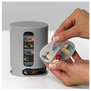 A person holds an open Mini Pill Storage Pill Box labeled MORNING in their left hand. This portable organizer fits into a gray circular holder and is divided by days from MON to SAT, with pills of various colors neatly arranged inside.