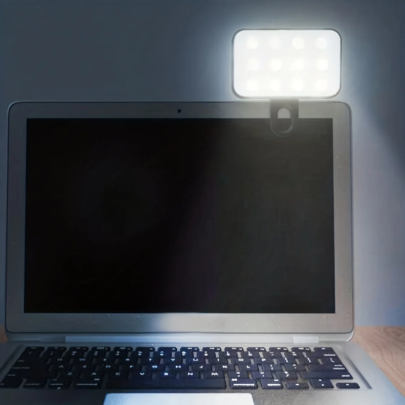 A Mini Selfie Fill Light with 3 color modes, adjustable brightness, and rechargeable design clips to the laptop’s top right, illuminating the keyboard and nearby area as it sits on a wooden surface.