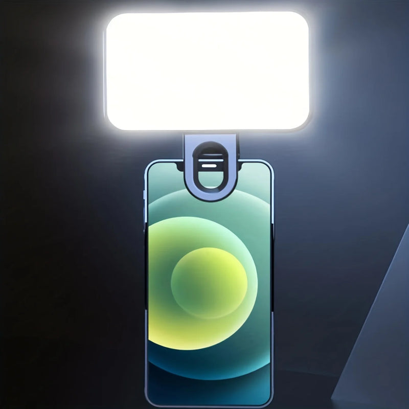 A smartphone has the Mini Selfie Fill Light clipped on top, its adjustable LED brightness and 3 color modes brightly illuminating the area ahead against a dark background.