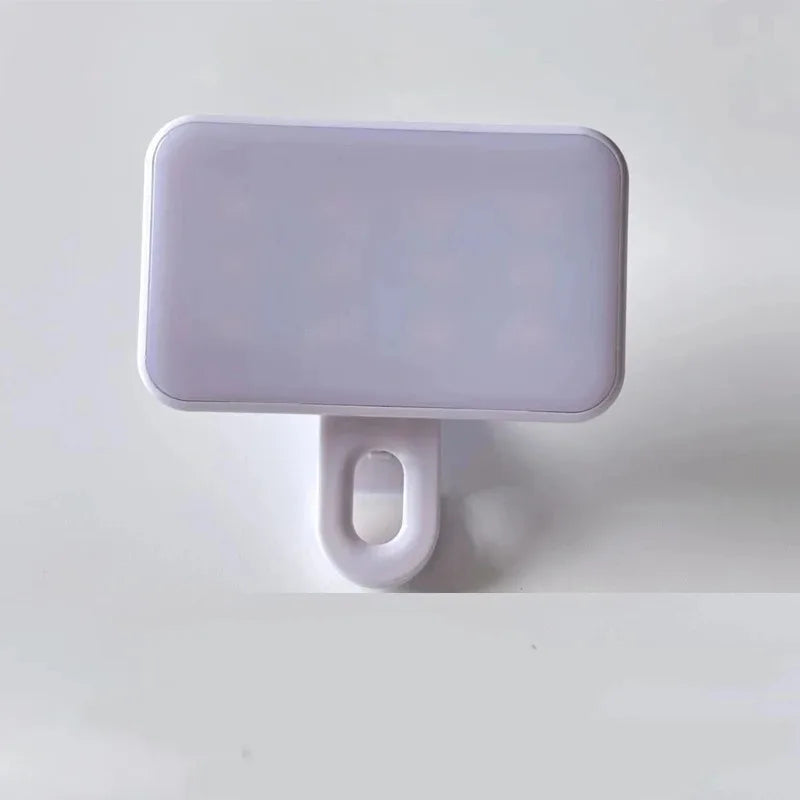 The Mini Selfie Fill Light features a rectangular white ABS body with rounded edges, a flat front, and is attached to a white base with an oval opening. It offers 3 color modes, adjustable brightness, and clips onto phones or computers for photography.