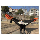A person in a face mask relaxes in an Automatic Quick-Opening Mosquito Net Hammock, set up in a grassy area. Houses and trees dot the background under a clear blue sky, creating an ideal pop-up for outdoor retreats.