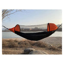An Automatic Quick-Opening Mosquito Net Hammock is elegantly suspended near a riverbank amidst dry grass and rocks, with distant buildings framed under a clear sky, ideal for a pop-up camping experience.