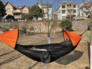 A person relaxes in a stylish Automatic Quick-Opening Mosquito Net Hammock, suspended between two trees in the park. Featuring black and orange with a mesh canopy for protection, residential houses peek through the greenery under a clear blue sky.