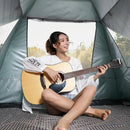 Seated cross-legged in an Automatic Tent Outdoor Camping Barbecue, a person plays an acoustic guitar. Theyre dressed in a white long-sleeve shirt and shorts, with a forest view visible through the tents waterproof opening in the background.