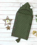 A green knitted baby sleeping bag shaped like a bear, featuring ears, side buttons, and a tail. Made from cozy cotton wool, it sits on a wooden surface with two decorative star ornaments and a small flower bouquet nearby.