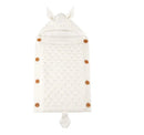 A cream-colored, knitted Baby Sleeping Bag with a hood, tiny ears, and a tail features a diamond pattern and wooden buttons. It is made with lamb cashmere lining and is displayed flat on a plain white background.