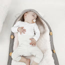 A baby sleeps peacefully in a cozy, knitted gray cocoon with button details, resembling a Baby sleeping bag. The infant wears a white outfit and has a pacifier, resting on a luxurious lamb cashmere-like lining.