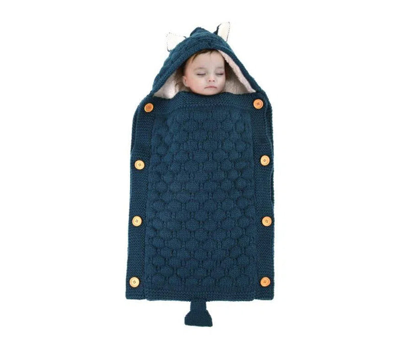 A baby sleeps peacefully in a blue knitted Baby Sleeping Bag with a hood featuring small ears and buttons. The lamb cashmere lining adds coziness, keeping the little one snug in the textured fabric.