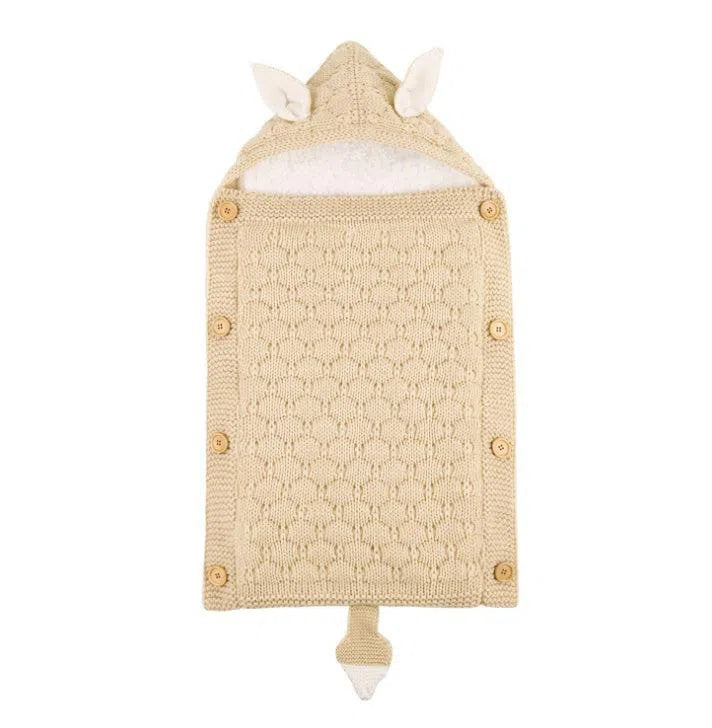 A beige knitted baby sleeping bag with a cozy lamb cashmere lining and hood featuring small animal ears. Wooden button closures on the sides complement its textured pattern, while a playful tail-like detail completes the design.