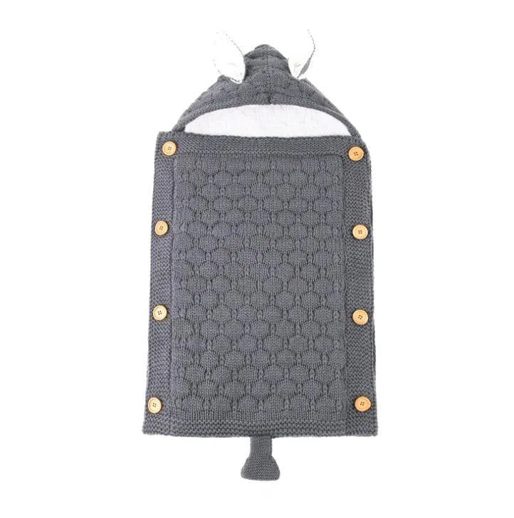 The Baby Sleeping Bag is a cozy grey knitted suit featuring a hood with ears and tail, designed to mimic an animal. It has a honeycomb pattern, wooden button closures on the sides, and is made from soft cotton wool for added comfort.