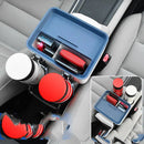 The Car Sundries Storage Cup Drink Mobile Phone Armrest Box Tissue Box, featuring a blue tray with slots for a red wallet, phone, and sunglasses, fits neatly between two front seats and includes cup holders for white and red cups. It complements the light gray fabric upholstery of the seats.