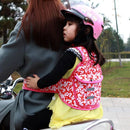 A child in a pink helmet and colorful vest hugs an adult in a grey coat from behind on a motorbike. The pair, traveling through a tree-lined area, use the Child safety rope motorcycle child safety belt for extra security.