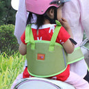 A child in a red shirt and pink helmet sits on a scooters back, secured by the green Child Safety Rope Motorcycle Child Safety Belt to someone in a striped shirt. Lush greenery unfolds behind, enhancing the serene scene.