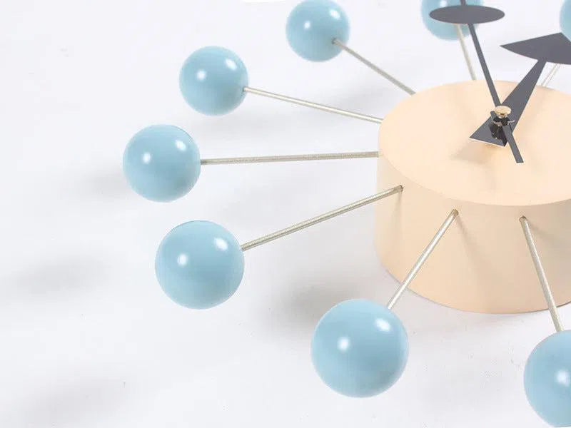 A close-up of the Clock, background wall, wall clock features light blue spheres on metal rods resembling molecules. Its minimalist beige face and black hands create a sleek focal point in contemporary interior design against a plain white background.