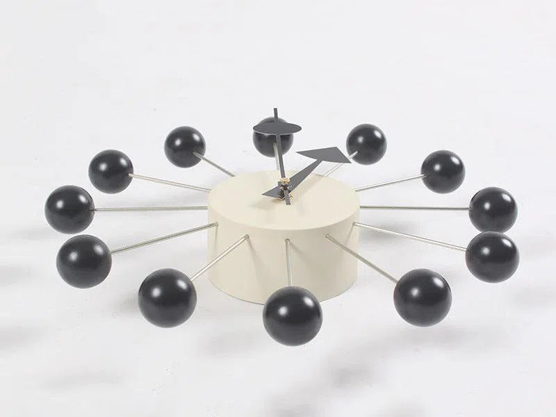 The Clock, background wall, wall clock features a modern European design with a cylindrical white base and twelve metal rods ending in black spheres for the hours. Minimalist black hour and minute hands enhance its striking appearance in any interior space.