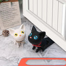 Two cute Door Handle Punch-free Door Stop cat stoppers, one black and one white with large eyes and collars, sit by a white cabinet door. A blurred text on paper is visible behind them. This charming scene effortlessly blends into the modern minimalist decor while providing anti-collision protection.