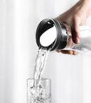 A hand pours water from a Glass Kettle Heat-resistant Cool Kettle with a shiny metal lid into a clear glass, both made of high borosilicate glass. The softly blurred background hints at a light, neutral setting.