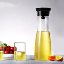 A tall glass carafe, the Glass Kettle Heat-resistant Cool Kettle Household Cold Kettle, made of high borosilicate glass holds yellow liquid beside a matching glass with an orange slice. Halved oranges are nearby, and cherry tomatoes and green leaves add color to the background.