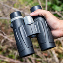A person holds a high-resolution binocular telescope featuring black textured grips and silver focus knobs for enhanced magnification, set against green foliage and dry branches.