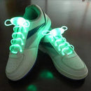 White sneakers feature LED Sport Shoe Laces with fiber-optic technology, glowing neon green on a dark surface. The looped laces emit bright light, giving the sneakers a futuristic look. Blue stripe details on the side add an extra pop of color to their sleek design.