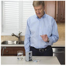 In a kitchen with wooden cabinets and a sink beneath a window, a man in a blue shirt holds a pill, standing beside the Mini Pill Storage Pill Box—a portable one-week distribution unit—alongside a small device, glass of water, and tray with more pills.