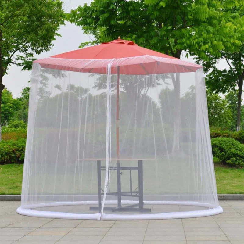 A red patio umbrella is surrounded by the Mosquito Net Outdoor Patio Umbrella Net Cover Roman Umbrella, making a perfect outdoor relaxation spot. Beneath, a square table and chair set beckons to leisure, all set on a paved area with lush green bushes and trees as the backdrop.
