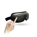 A hand approaches a button on the New Eye Spa And Music Therapy mask, a sleek black design with graphene technology. Its adjustable strap suits an at-home experience. Against a white background, it seems to float in air.