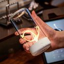 A hand holds the New LED Camping Light, a stylish glass lamp with a white base resembling a portable night light. It features an artistic filament glow for warmth and charm, with an electronic device in the background hinting at its Type-C rechargeable convenience.