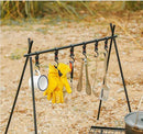 A foldable Outdoor Camping Aluminum Alloy Rack in sandy terrain holds a mug, yellow gloves, ladle, tongs, slotted spoon, and spatula. Grass and a cooking pot appear in the background.