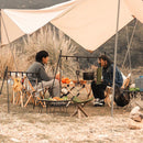 Two people converse under a large tent at a campsite, seated on foldable chairs around a small table with camping gear and food, including oranges. Nearby stands the Outdoor Camping Aluminum Alloy Large Foldable Portable Clothes Drying Rack amidst the tall grass and woods.