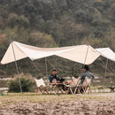 Two people are camping by the river under a white tarp tent, surrounded by lush hills. Seated on folding chairs at a table with camping gear, they have an Outdoor Camping Aluminum Alloy Large Foldable Portable Clothes Drying Rack nearby, perfectly suited for their scenic adventure.