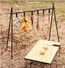 A triangular, portable aluminum-alloy shelf holds utensils, gloves, and a mug on hooks. Below, an outdoor scene shows a wooden table with a knife and two peeled potatoes resting on dirt. Product: Outdoor Camping Aluminum Alloy Large Foldable Portable Clothes Drying Rack For Camping Travel Triangle.