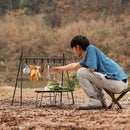 At an outdoor campsite, a person in a blue shirt and gray pants crouches on a stool, holding a spoon near an Outdoor Camping Aluminum Alloy Large Foldable Portable Clothes Drying Rack. Utensils, gloves, and a pan hang from it. A small table with chopped vegetables is in front of them amidst the trees.