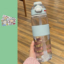 A hand holds the Portable Large Capacity Plastic Cup, a temperature-resistant clear bottle featuring a light blue grip and lid with a straw. Its pictured on a wooden floor with green decor adorned by colorful cartoon character stickers.