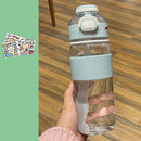 A hand holds the Portable Large Capacity Plastic Cup with a transparent design, light blue lid and grip. The warm wooden floor complements two cute character sticker sheets on green, meeting your hydration needs stylishly.