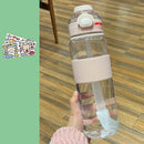 A hand holds a Portable Large Capacity Plastic Cup with a pink lid and ribbed grip on a wooden floor, ideal for hydration. Decorative stickers add charm to the green background on the left.
