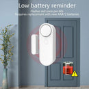 The white Smart Home Door And Window Sensing Alarm on the closed blue door flashes a red low battery reminder every 60 seconds, indicating its time to replace two AAA batteries. A potted plant is on the left, and an image shows a warning icon. Enhance your home security system today.