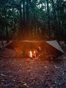 A person sits beside a campfire under the Sunscreen And Rainproof Camping Barbecue Leisure Awning in the dense forest. At dusk, the fire lights up their tarp haven, surrounded by towering trees and scattered leaves.