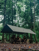 A small group sits under a Sunscreen And Rainproof Camping Barbecue Leisure Awning in a forest, surrounded by trees. They gather around a table with a lantern, seated on white and brown camping chairs. Leaf-covered ground suggests wetness.