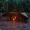 In a dimly lit forest, someone tends to a small campfire under the Sunscreen And Rainproof Camping Barbecue Leisure Awning. A lantern sits on a nearby wooden bench, hinting at evening or early morning in the dense woods.
