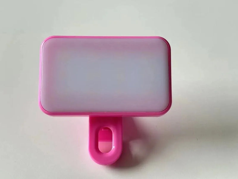 The Mini Selfie Fill Light, featuring 2835 LED beads, a pink frame, and a clip, offers 3 color modes and adjustable brightness. Shown on a plain white surface, it's rechargeable and clips onto phones or computers for photography lighting.